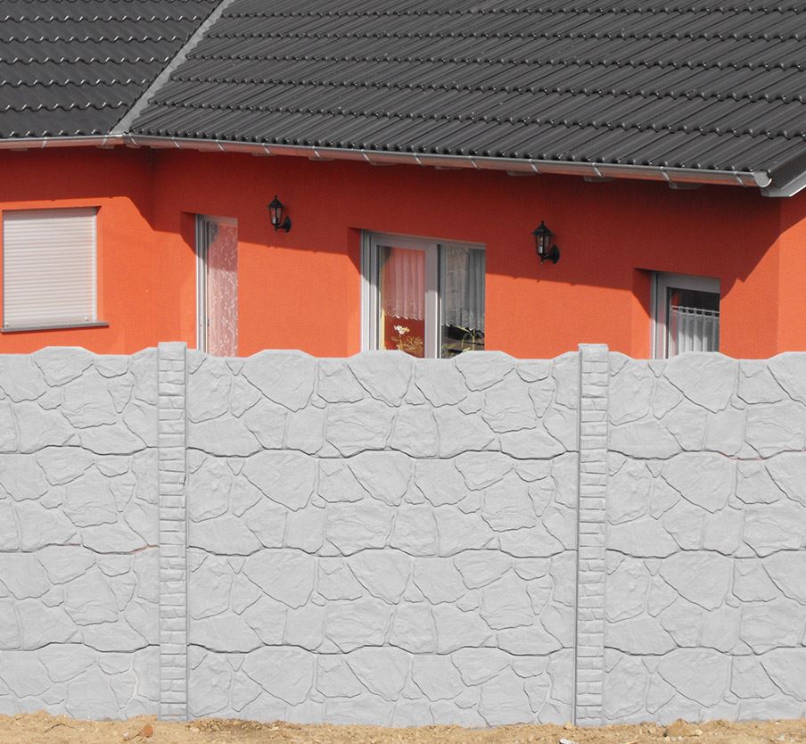 Ein grauer Betonzaun mit Felsoptik im Vordergrund, dahinter ein orangefarbenes Haus mit dunklem Ziegeldach. Der Zaun hat eine raue, steinähnliche Textur und verdeckt teilweise die Fassade. Am Haus sind Fenster und Außenbeleuchtung zu sehen. Der Kontrast zwischen dem grauen Zaun und der leuchtend orangen Hauswand ist auffällig."