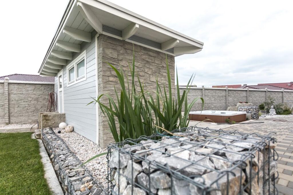 34 | Moderner Garten mit Betonzaun und Nebengebäude. Steinoptik-Betonplatten umgeben den Bereich. Im Vordergrund ein Gabione mit Pflanzen, im Hintergrund ein Whirlpool. Helle Farbtöne und gepflegte Landschaftsgestaltung vermitteln einen eleganten Eindruck