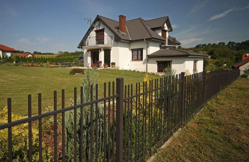 Weißes Einfamilienhaus mit grauem Dach, großem Rasen und schwarzem Metallzaun im Vordergrund