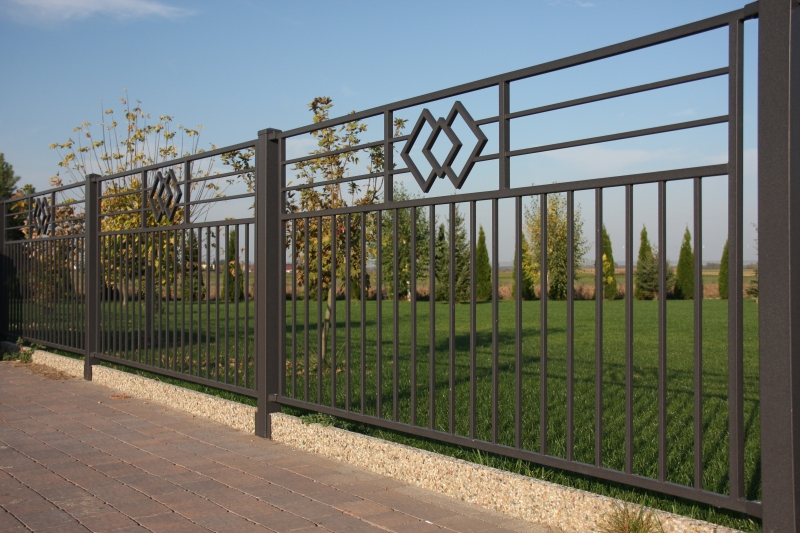 Schwarzer Metallzaun mit dekorativem Rautenmuster vor einem grünen Garten mit Nadelbäumen im Hintergrund