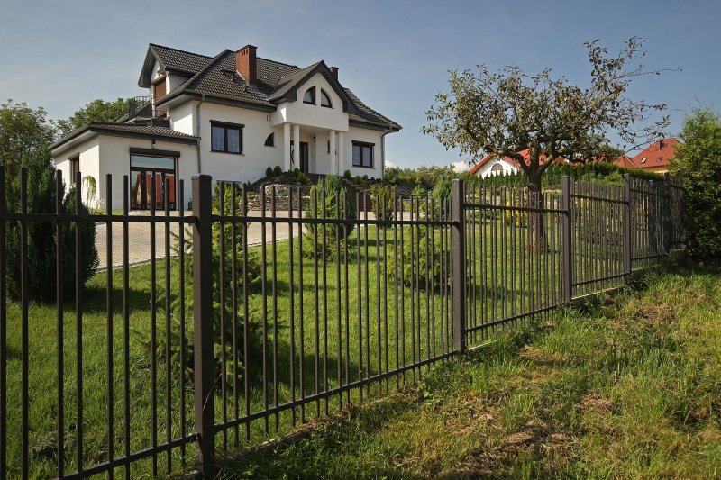 Weißes Einfamilienhaus mit grauem Dach hinter einem schwarzen Metallzaun, umgeben von gepflegtem Rasen und Bäumen