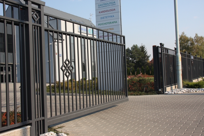 Schwarzes Metallzauntor mit dekorativem Rautenmuster vor einem Medizinzentrum mit Praxisschildern für verschiedene Fachärzte
