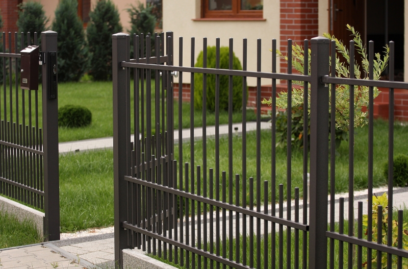 Schwarzer Metallzaun mit vertikalen Stäben vor einem gepflegten Garten mit grünem Rasen und Gehweg