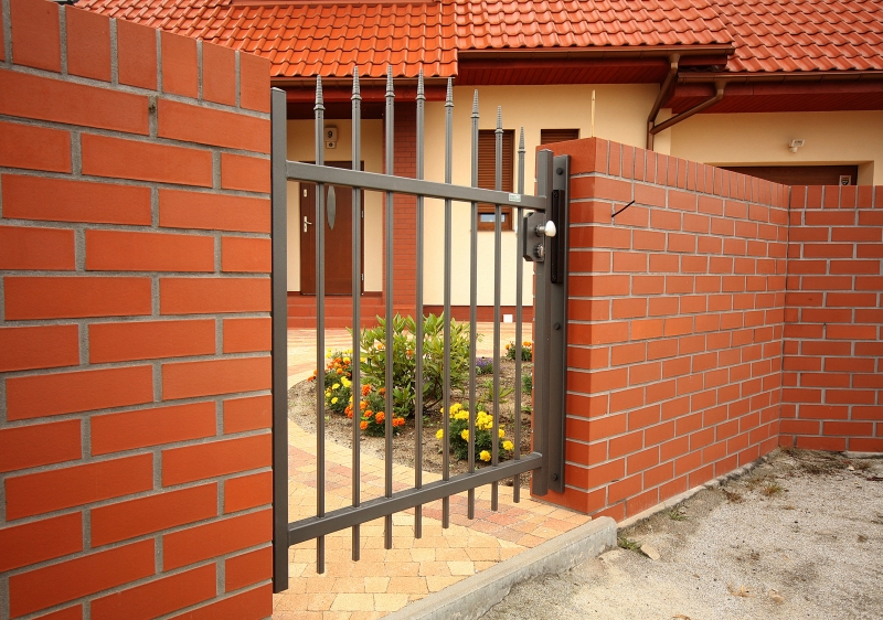 Schwarzes Metallgartentor zwischen roten Ziegelmauern, davor ein kleines Blumenbeet mit gelben und orangen Blüten