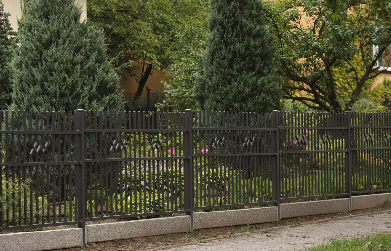 Eleganter schwarzer Metallzaun mit dekorativem Muster vor hohen Koniferen und blühenden Gartenpflanzen