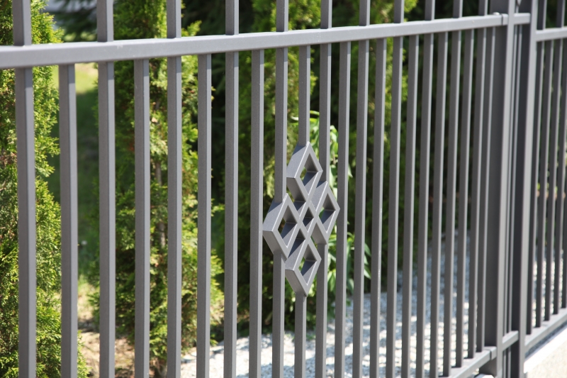 Grauer Metallzaun mit dekorativem Rautenmuster, im Hintergrund grüne Gartenvegetation