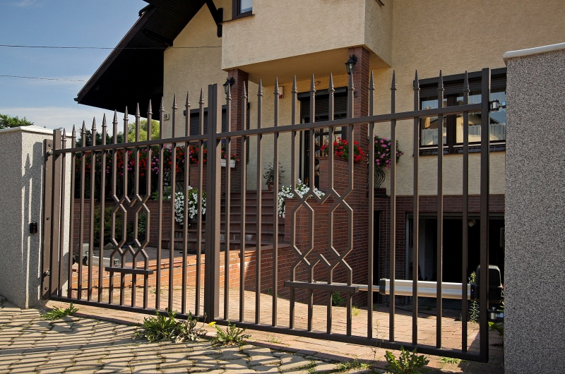 Eleganter Metallzaun vor einem beigen Haus mit roten Blumen, geometrische Muster im Zaun integriert