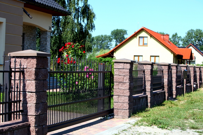 Eleganter schwarzer Metallzaun zwischen rosa Steinsäulen vor einem Haus mit rotem Dach, geschmückt mit roten Blumen