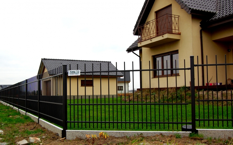 Gelbes Einfamilienhaus mit Balkon hinter einem schwarzen Metallzaun, gepflegter Rasen im Vordergrund