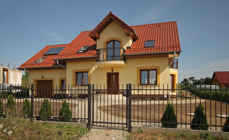 Gelbes Einfamilienhaus mit rotem Ziegeldach, Balkon und Solarplatten, umgeben von schwarzem Metallzaun