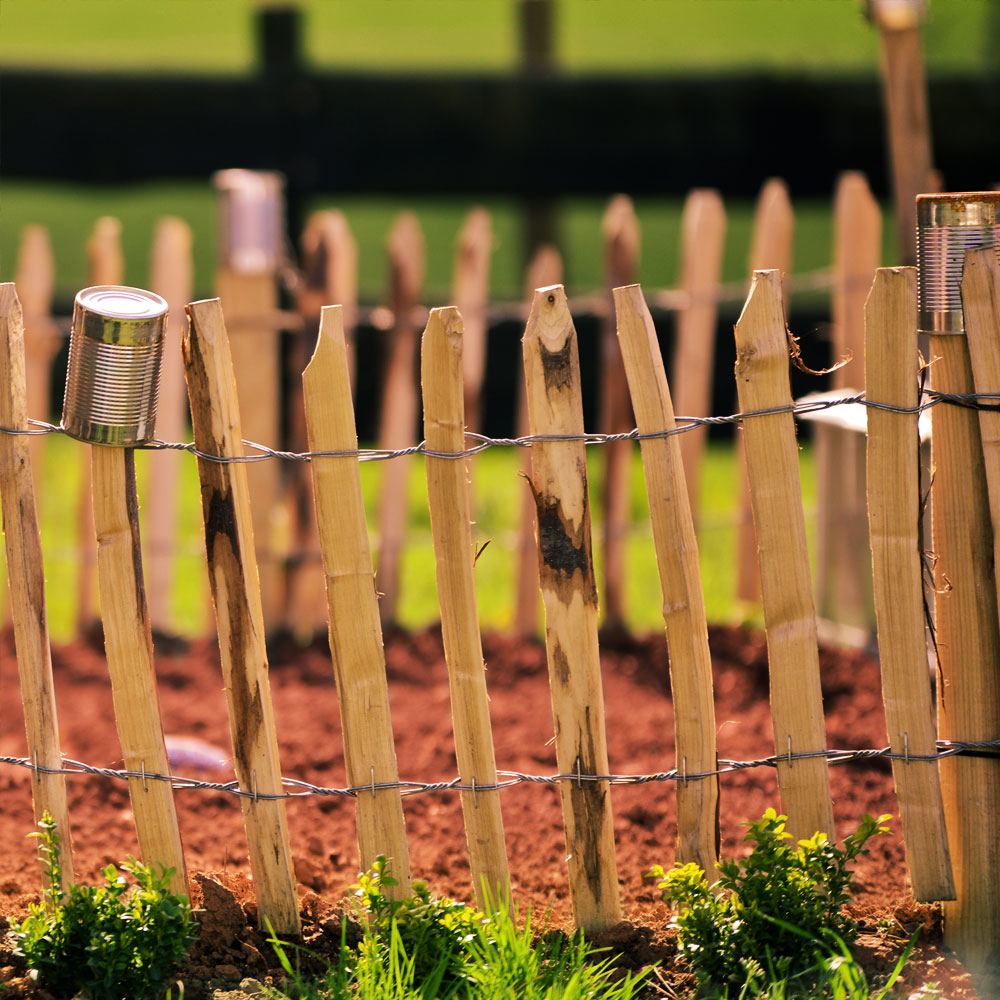 Selbstgebauter Gartenzaun aus Haselnussmit Stacheldraht, Blechdosen als Dekoration auf rotem Erdboden