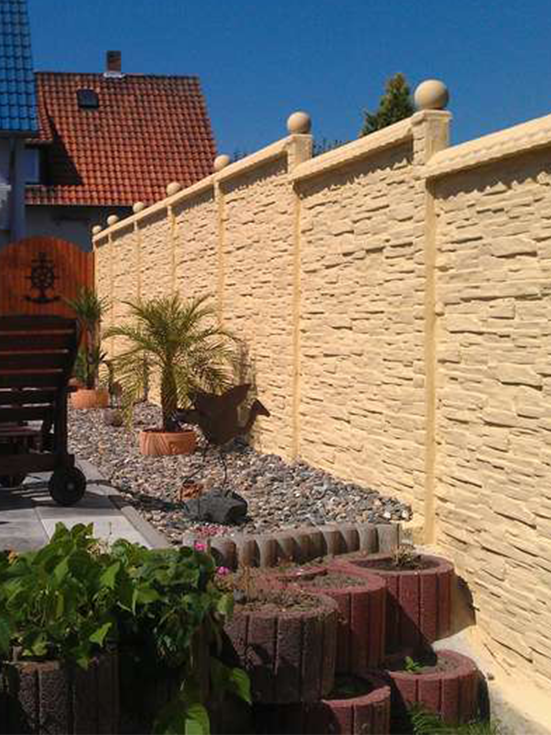 Sandsteinfarbener Betonzaun mit strukturierter Oberfläche in einem gepflegten Garten. Der Zaun hat dekorative Säulen mit runden Abschlüssen. Davor befindet sich ein geschwungenes Blumenbeet mit roten Ziegeln und verschiedenen Pflanzen, einschließlich einer kleinen Palme. Im Hintergrund ist ein Haus mit rotem Ziegeldach zu sehen